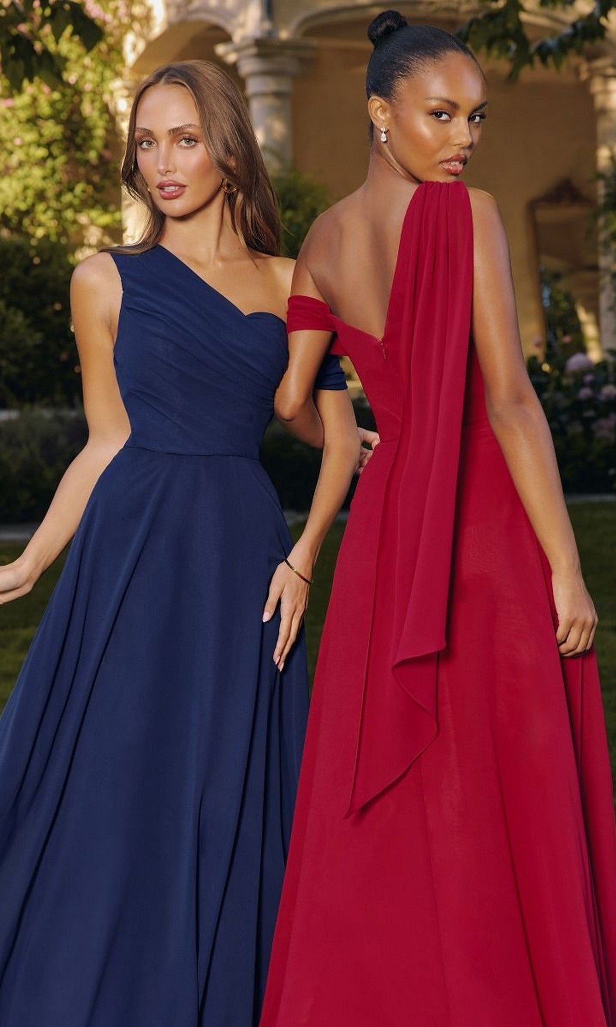 Two women stand outdoors in elegant gowns; one wears a navy blue one-shoulder dress, while the other stuns in the red Amoris T716 Asymmetric Long A-Line Prom Dress featuring a beautifully draped detail.